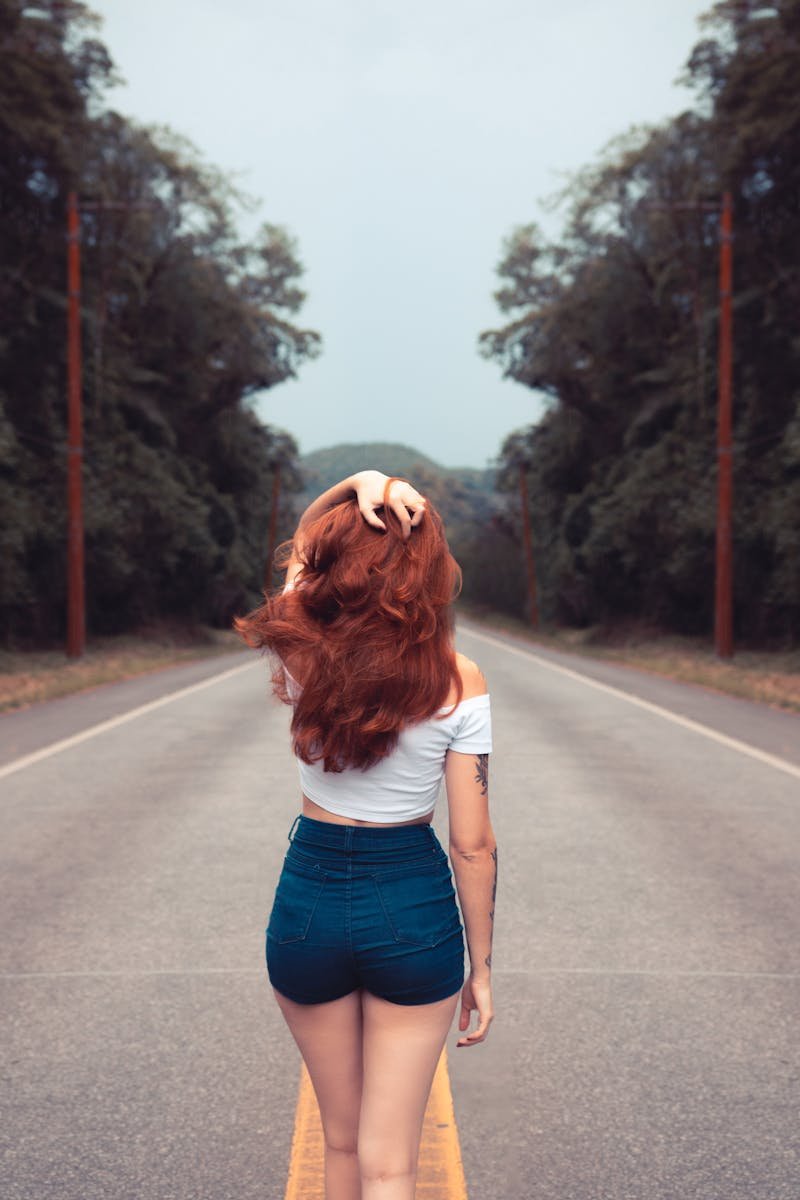 Red-haired woman in jean shorts walking down a forest road, showcasing style and freedom.